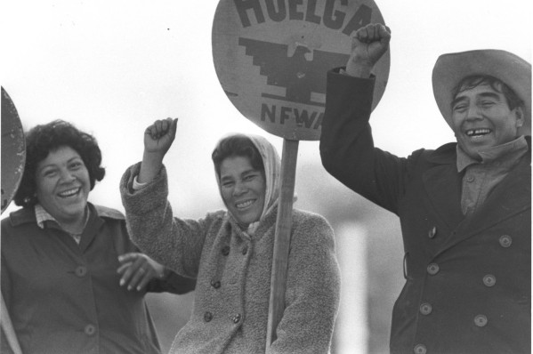 Photographic print of three smiling people raising their fists outdoors, one holding a circular NFWA "Huelga" strike sign