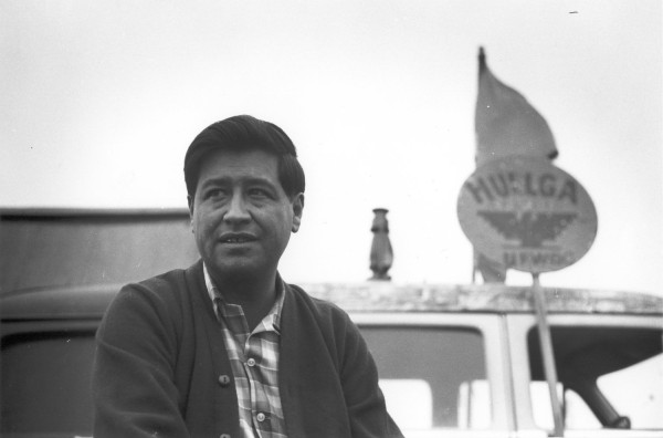 Photographic print of Cesar Chavez seated in front of a worn vehicle topped with a lantern and a circular 'Huelga' sign with a flag
