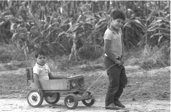 Photographic print of a smiling boy pulling a rope attached to a wooden wagon, in which a smaller toddler sits, on a dirt path before a field
