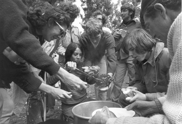 Photographic print of a group of young people crowding around a large metal bowl outdoors, serving and eating food with trees in the background