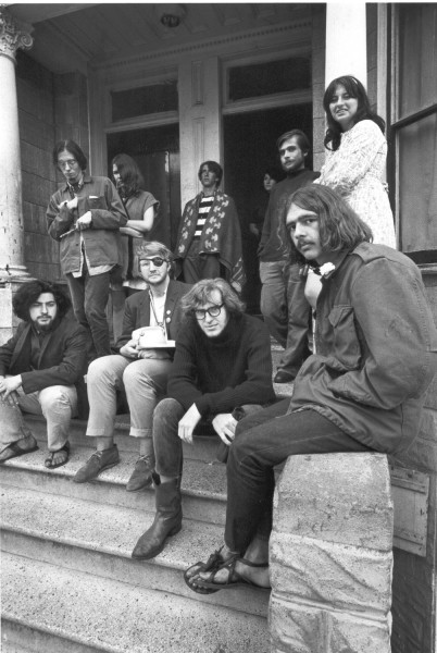 Photographic print of a group of long-haired men and women gathered on the front steps and porch of a house, wearing casual 1960s clothing