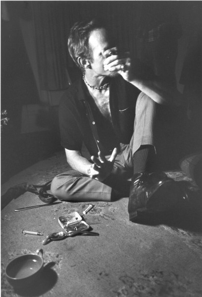 Photographic print of a man seated cross-legged on a floor, holding an object to his face. Hypodermic needles, a spoon, and a small case rest on the floor nearby