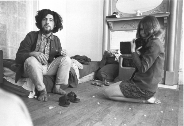Photographic print of a bearded man seated on a mattress and a woman kneeling on a bare wooden floor, both barefoot in a cluttered room with a fireplace