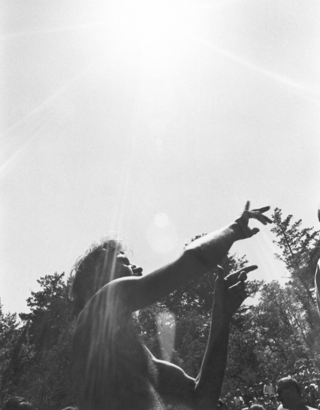 Photographic print showing a silhouetted person with curly hair reaching both arms upward toward a bright sun. Trees and a crowd are visible in the background below