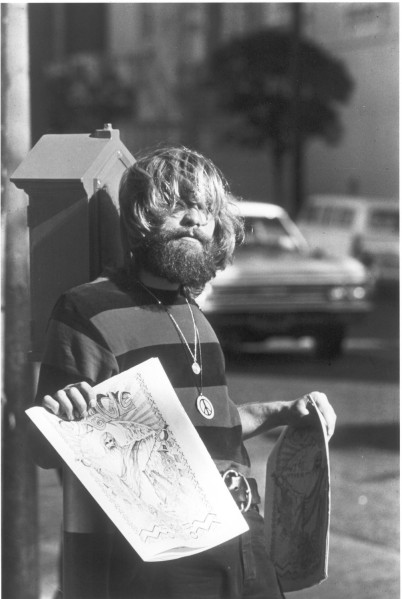 Photographic print of a bearded, long-haired person wearing a peace symbol pendant, holding detailed pen drawings on a city street