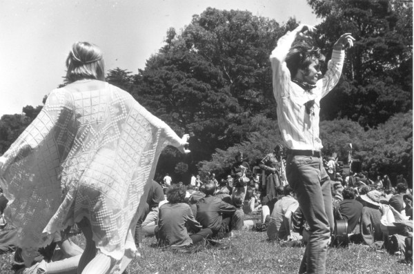 Photographic print of two young people dancing on a grass lawn, one draped in a patterned shawl, arms raised. A large crowd sits on the ground beneath tall trees in the background