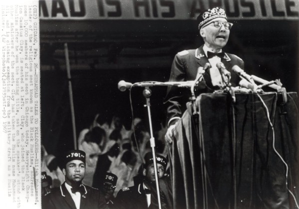 Photographic print of Elijah Muhammad speaking at a podium with multiple microphones, wearing a crown, as Muhammad Ali and other men in FOI caps sit on stage behind him
