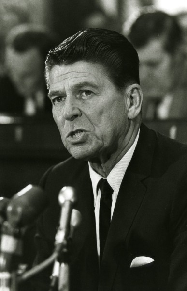 Photographic print of Governor Ronald Reagan speaking at a press conference, wearing a dark suit and tie, with multiple microphones in the foreground