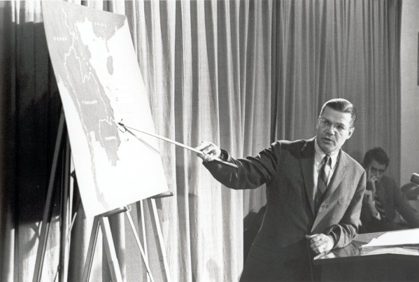 Photographic print of Secretary of Defense Robert S. McNamara using a pointer at a press conference, gesturing toward a large map of Southeast Asia on an easel