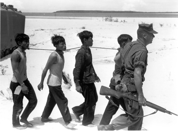 Photographic print of an armed Marine leading four prisoners tied together by a rope around their necks across a sandy open terrain