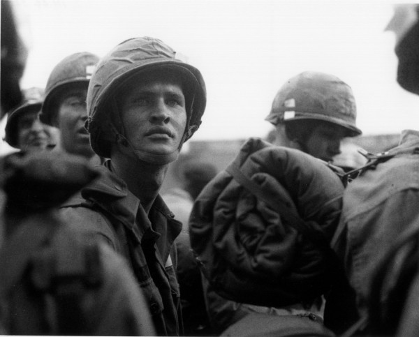 Photographic print of soldiers in helmets and field gear looking upward, crowded together in a close group against an overcast sky