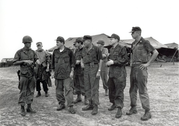 Photographic print of a group of military personnel in combat uniforms standing on open ground, with large canvas tents visible in the background