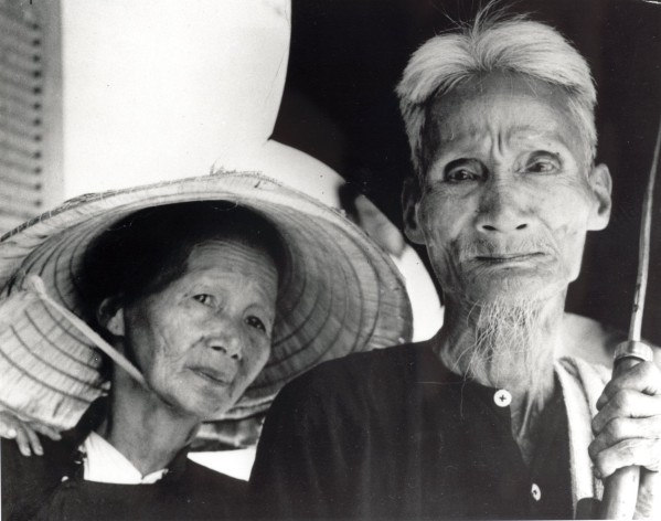 Photographic print of an elderly woman wearing a conical straw hat and an elderly man with a white beard holding a cane, both with deeply lined faces