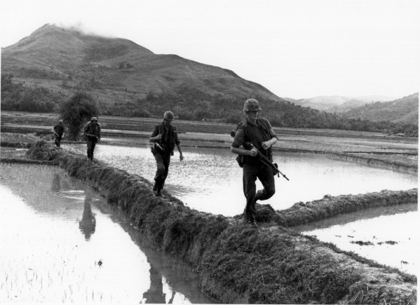 Photographic print of four armed, helmeted Marines moving single-file along narrow rice paddy dikes, with flooded fields and mountains in the background