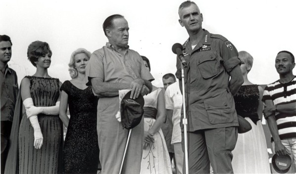 Photographic print of General Westmoreland in U.S. Army uniform speaking at a microphone beside Bob Hope, who holds a golf club, with a group of performers behind them