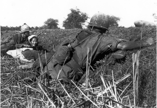 Photographic print of a helmeted soldier lying prone in a field, arm extended toward a woman and child crouching low among dense vegetation and brush