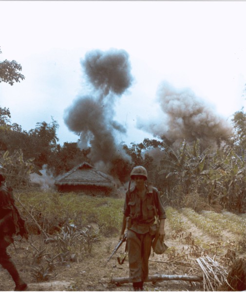 Photographic print of a U.S. Marine in helmet and combat gear walking forward as two large columns of dark smoke rise behind a thatched structure amid dense jungle vegetation