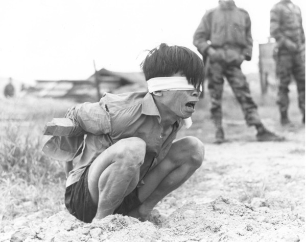 Photographic print of a blindfolded, bound prisoner crouching on bare ground, mouth open. Two soldiers in camouflage stand in the background