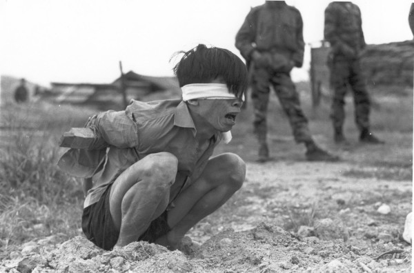 Photographic print of a blindfolded, bound prisoner squatting on bare ground and crying, with soldiers in camouflage fatigues standing in the background