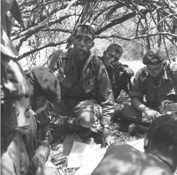 Photographic print of soldiers in military fatigues crouched under tree branches, gathered around a map on the ground during a field briefing