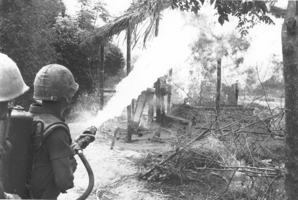 Photographic print of two U.S. Marines in helmets directing a flamethrower at a burning thatched structure surrounded by trees and debris