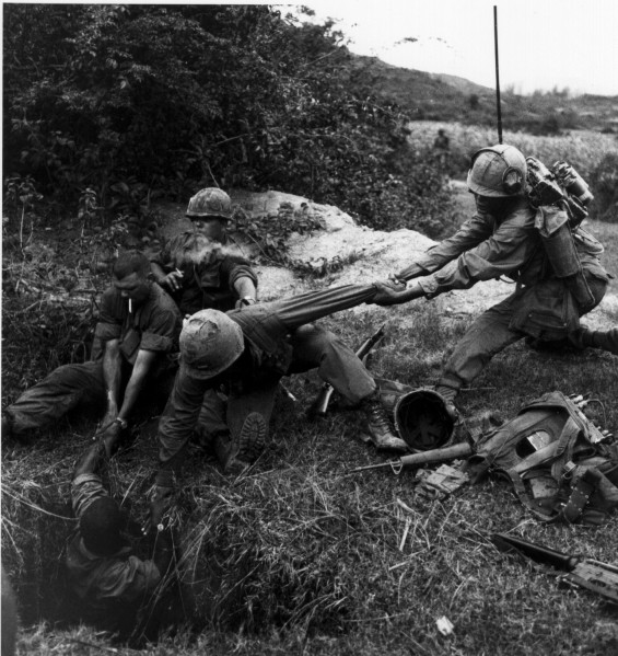 Photographic print of three helmeted soldiers lowering a fourth soldier into a ground tunnel, with gear and weapons scattered on the grass nearby