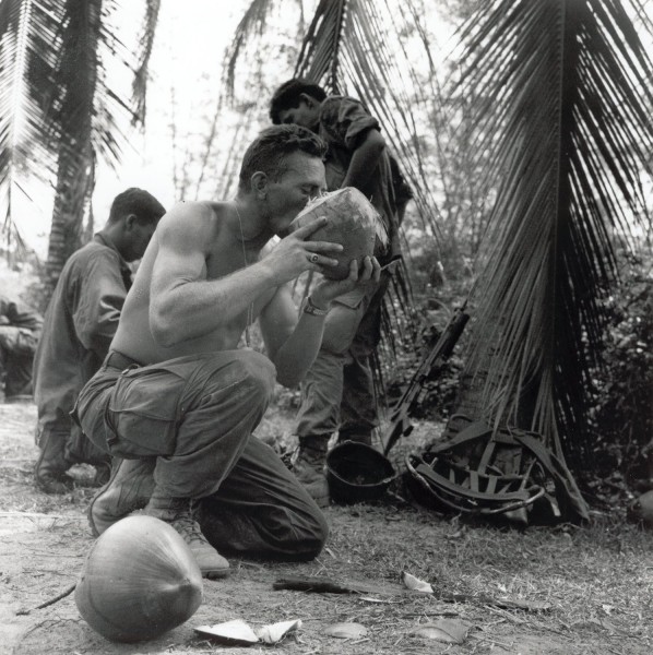 Photographic print of a shirtless soldier kneeling and drinking from a coconut beneath palm trees, with other soldiers, gear, and a rifle visible nearby