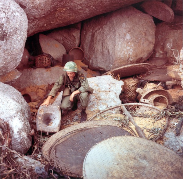 Photographic print of a helmeted soldier in olive fatigues crouching among large boulders, examining scattered woven baskets and debris in a rocky cave