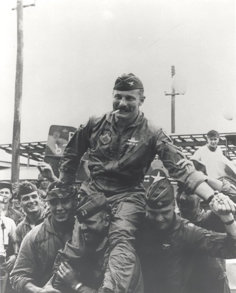 Photographic print showing Colonel Robin Olds in flight suit and garrison cap, carried on the shoulders of smiling fellow pilots near an aircraft on the flight line