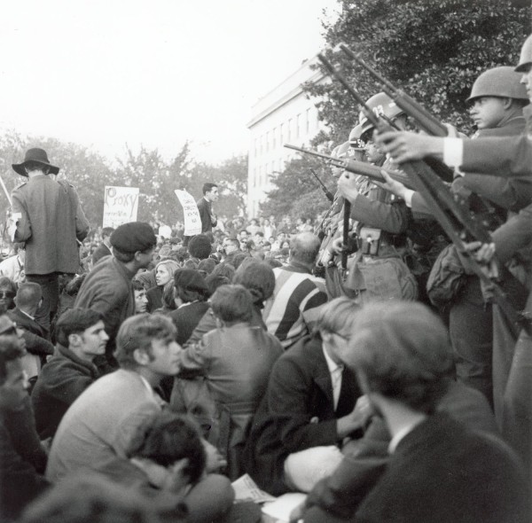 Photographic print of a dense crowd of seated protesters facing a line of helmeted military police holding rifles, with protest signs visible in the background