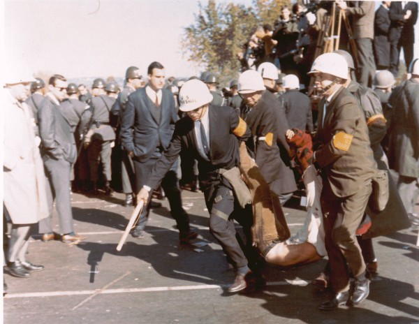 Photographic print of U.S. marshals in white helmets and armbands dragging a limp protester across pavement, surrounded by a large crowd