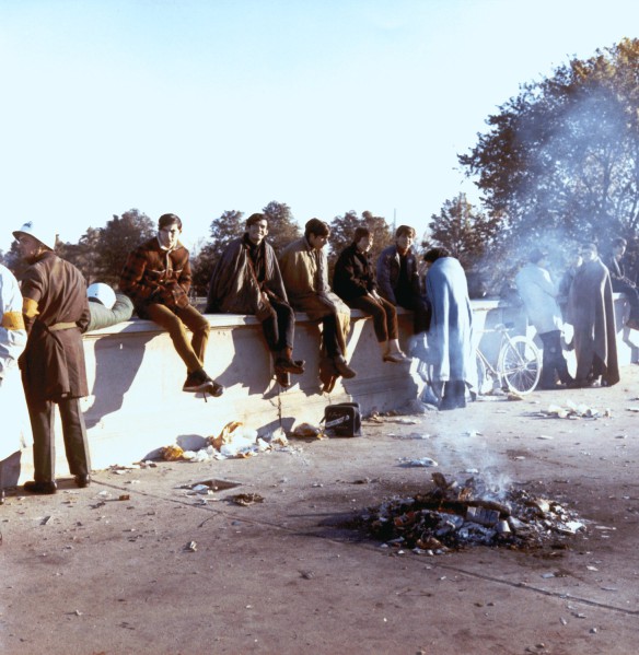 Photographic print of several people seated on a low concrete wall near a smoldering bonfire on a littered plaza, with trees and a bicycle visible behind them