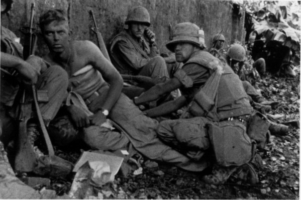 Photographic print of several combat-equipped Marines resting against a wall, one shirtless with bandaging visible on his torso, others in helmets and field gear