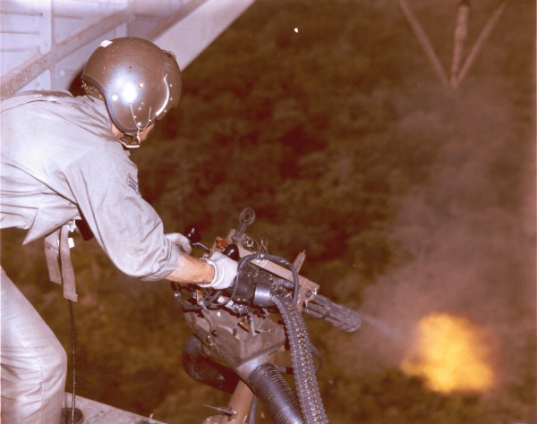 Photographic print of a U.S. Air Force HH-3 crewman in flight suit and helmet operating a mounted machine gun. Muzzle flash and smoke are visible at the gun barrel with dense forest canopy below