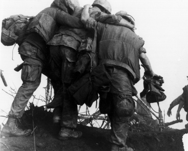 Photographic print of two Marines in combat gear supporting a wounded Marine between them, moving across rough terrain with sparse vegetation