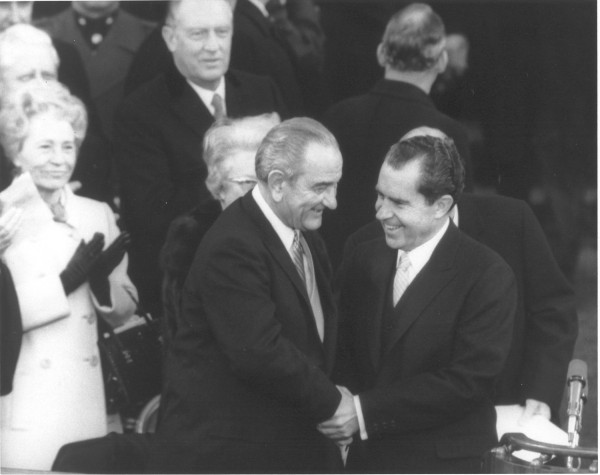 Photographic print of President Johnson and president-elect Nixon shaking hands outdoors, both in dark suits, surrounded by onlookers in formal attire