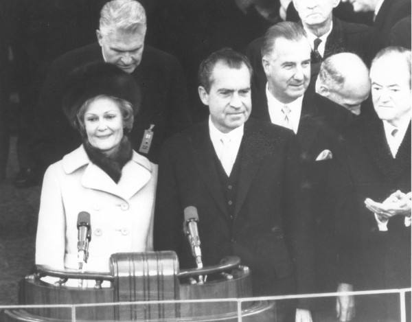 Photographic print of Pat Nixon in a light coat standing at a podium with microphones beside Richard Nixon in a dark suit, surrounded by a crowd of formally dressed figures