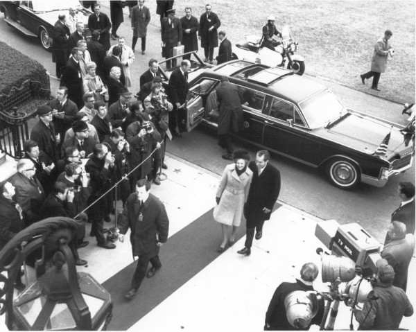 Photographic print of President Richard Nixon and Pat Nixon walking beside a large black limousine, surrounded by press photographers and a TV pool camera