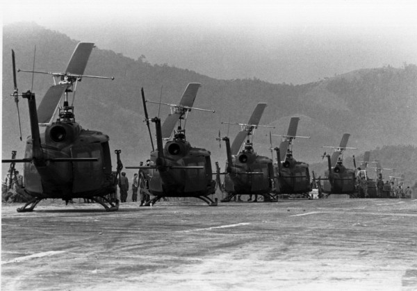 Photographic print of a row of military helicopters parked on a landing pad, with personnel nearby and forested mountains in the background