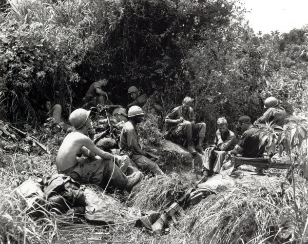 Photographic print of a group of helmeted soldiers resting on a hillside amid dense jungle vegetation, rifles and equipment visible nearby