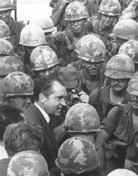 Photographic print of President Nixon in a suit, surrounded by a large group of soldiers wearing camouflage helmets and fatigues, smiling and shaking hands