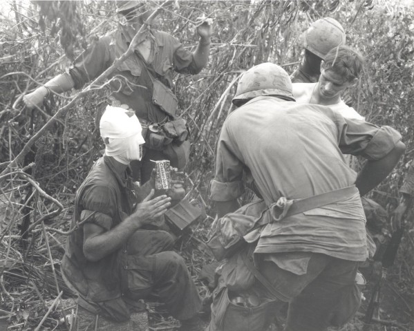 Photographic print of several soldiers in military gear gathered in dense brush. One soldier has his head heavily bandaged and holds a Cracker Jack box while others lean in around him