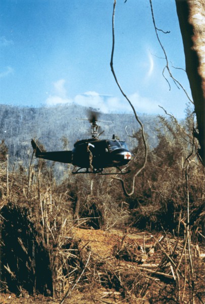 Photographic print of a UH-1D Medevac helicopter marked with a red cross hovering low over a cleared, debris-strewn area surrounded by sparse trees and hills