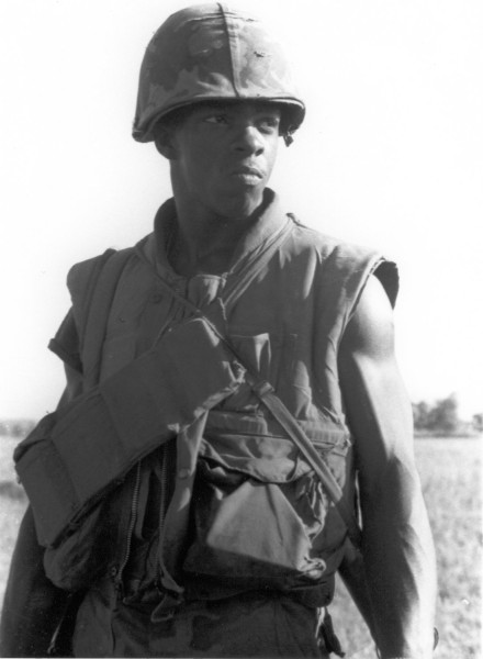 Photographic print of a Black marine in a camouflage helmet and flak vest with gear straps, standing in an open field with trees in the background