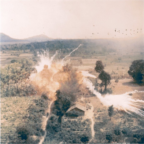 Photographic print taken from the air showing napalm bombs exploding in fireballs and smoke over structures amid trees and fields, with mountains in the distance