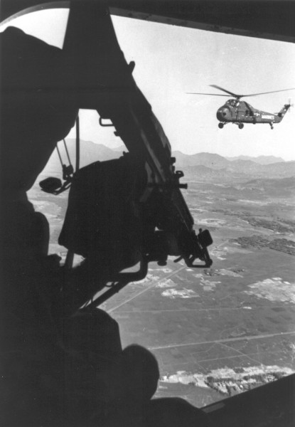 Photographic print taken from inside an aircraft showing a silhouetted soldier aiming a gun toward a Marines helicopter flying over a mountainous landscape