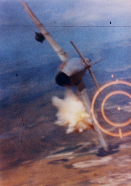 Photographic print of a MiG fighter aircraft banking steeply, with a burst of flames and smoke erupting near the left wing and fuselage. A gunsight reticle is visible at right