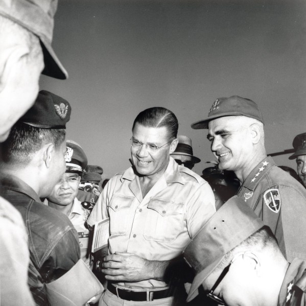 Photographic print of McNamara and General Westmoreland in khaki uniforms conversing with military officers outdoors, surrounded by a group of personnel