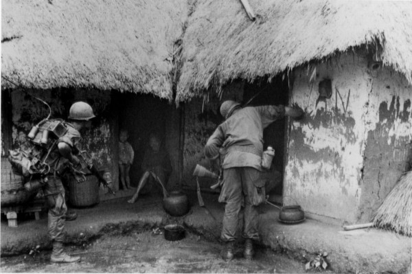 Photographic print of two helmeted soldiers with full gear examining the exterior and doorway of thatched-roof mud structures, with a child visible in the doorway