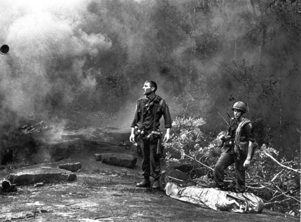 Photographic print of two uniformed soldiers standing in a jungle clearing amid smoke and debris, with a body wrapped in a blanket on the ground nearby